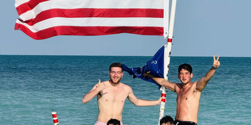 Four shirtless men in swim trunks are standing in the ocean water, with two men sitting on the shoulders of the other two. They are holding a large American flag on a pole, which is waving in the wind. The sky is clear and blue, and the water is calm. The men appear to be enjoying themselves, with one making a shaka hand sign and another flashing a peace sign.