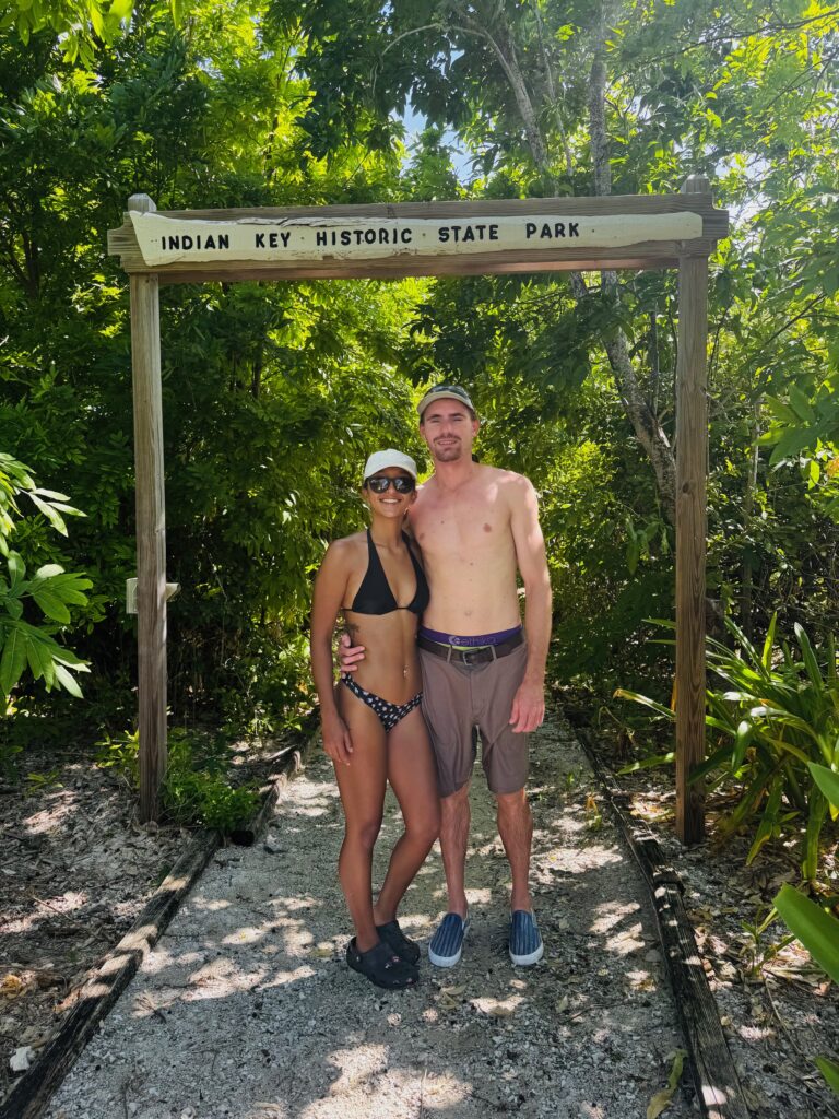 A couple stands on a gravel path under a wooden sign that reads "Indian Key Historic State Park." The woman is wearing a black bikini, black Crocs, a white cap, and sunglasses. The man is shirtless, wearing brown shorts, a belt, a cap, and slip-on shoes. They are surrounded by lush green foliage.