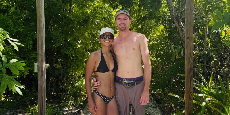 A couple stands on a gravel path under a wooden sign that reads "Indian Key Historic State Park." The woman is wearing a black bikini, black Crocs, a white cap, and sunglasses. The man is shirtless, wearing brown shorts, a belt, a cap, and slip-on shoes. They are surrounded by lush green foliage.