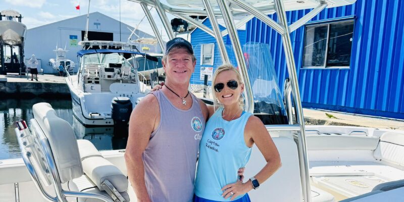 A man and a woman standing together on a white 248 Commander boat. The man is wearing a gray tank top, a cap, and colorful shorts, while the woman is wearing sunglasses, a light blue sleeveless top with "Captain Rebekah" written on it, and a blue skirt. The boat is docked near a blue building with other boats visible in the background.