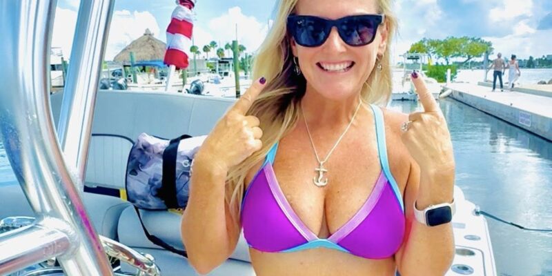 A woman wearing a purple and blue bikini and black sunglasses is standing on a boat. She is smiling and pointing at her face with both hands. She has blonde hair, an anchor necklace, and a smartwatch on her left wrist. The background shows a marina with boats, a dock, and a partly cloudy sky.