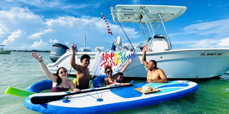 Four people are sitting and standing on a large inflatable paddleboard in shallow water near a white motorboat. They are smiling and waving, with a "Happy Birthday" banner displayed on the boat behind them. A small birthday cake is placed on the paddleboard. The sky is bright blue with some clouds.