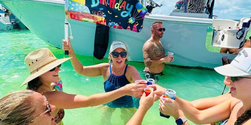 A group of people celebrating in shallow, clear green water near a white boat. One woman is holding a colorful "Happy Birthday" flag. The group is toasting with small cups and cans, enjoying a sunny day with blue skies. Some are wearing swimsuits, sunglasses, hats, and caps. The boat has the label "268 Commander" on its roof.
