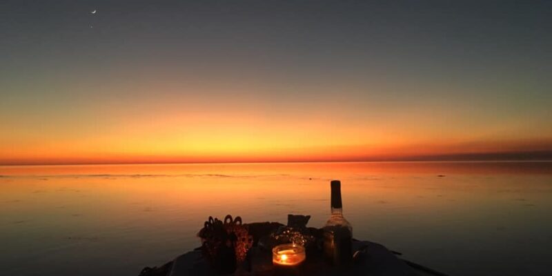 A calm sunset over a body of water with a gradient sky transitioning from deep blue to orange near the horizon. In the foreground, the bow of a boat is visible with a lit candle, a bottle, and some decorative items placed on it. A crescent moon is faintly visible in the upper left part of the sky.