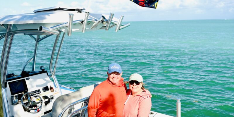 A couple standing on a white boat in the water, smiling and embracing. The man is wearing a blue cap, orange long-sleeve shirt, and blue shorts with a pineapple pattern. The woman is wearing a light green cap, sunglasses, a pink hoodie, and black shorts. A colorful flag with "HAPPY ANNIVERSARY" in bold yellow and red letters is flying above them. The background shows calm blue-green water and a clear sky with a few clouds.