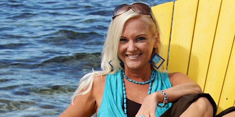 A woman with long blonde hair is sitting on a yellow Adirondack chair by the water. She is wearing a turquoise sleeveless top, brown pants, large blue geometric earrings, a turquoise necklace, a large decorative ring, and sunglasses on her head. She is smiling and appears relaxed with one leg bent and her arm resting on the chair. The background shows a body of water and a wooden pier.