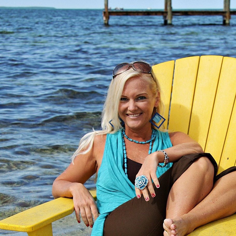 A woman with long blonde hair is sitting on a yellow Adirondack chair by the water. She is wearing a turquoise sleeveless top, brown pants, large blue geometric earrings, a turquoise necklace, a large decorative ring, and sunglasses on her head. She is smiling and appears relaxed with one leg bent and her arm resting on the chair. The background shows a body of water and a wooden pier.
